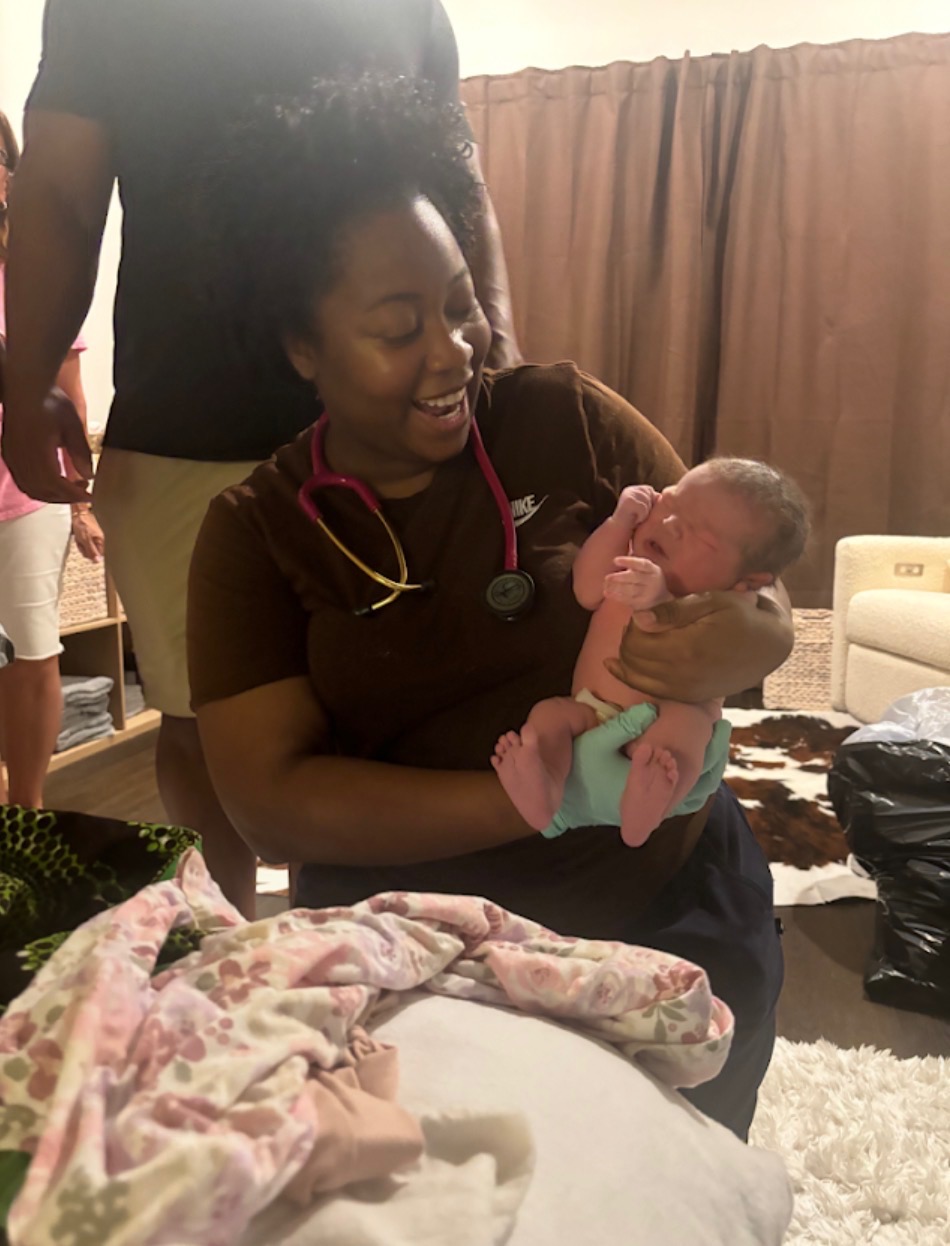 A birth worker in scrubs smiling as she holds a newborn at a home birth
