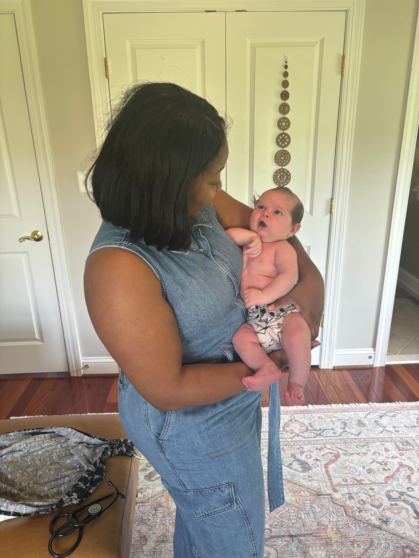 Jael standing in a sunlit bedroom holding a wide eyed newborn during a postpartum home visit