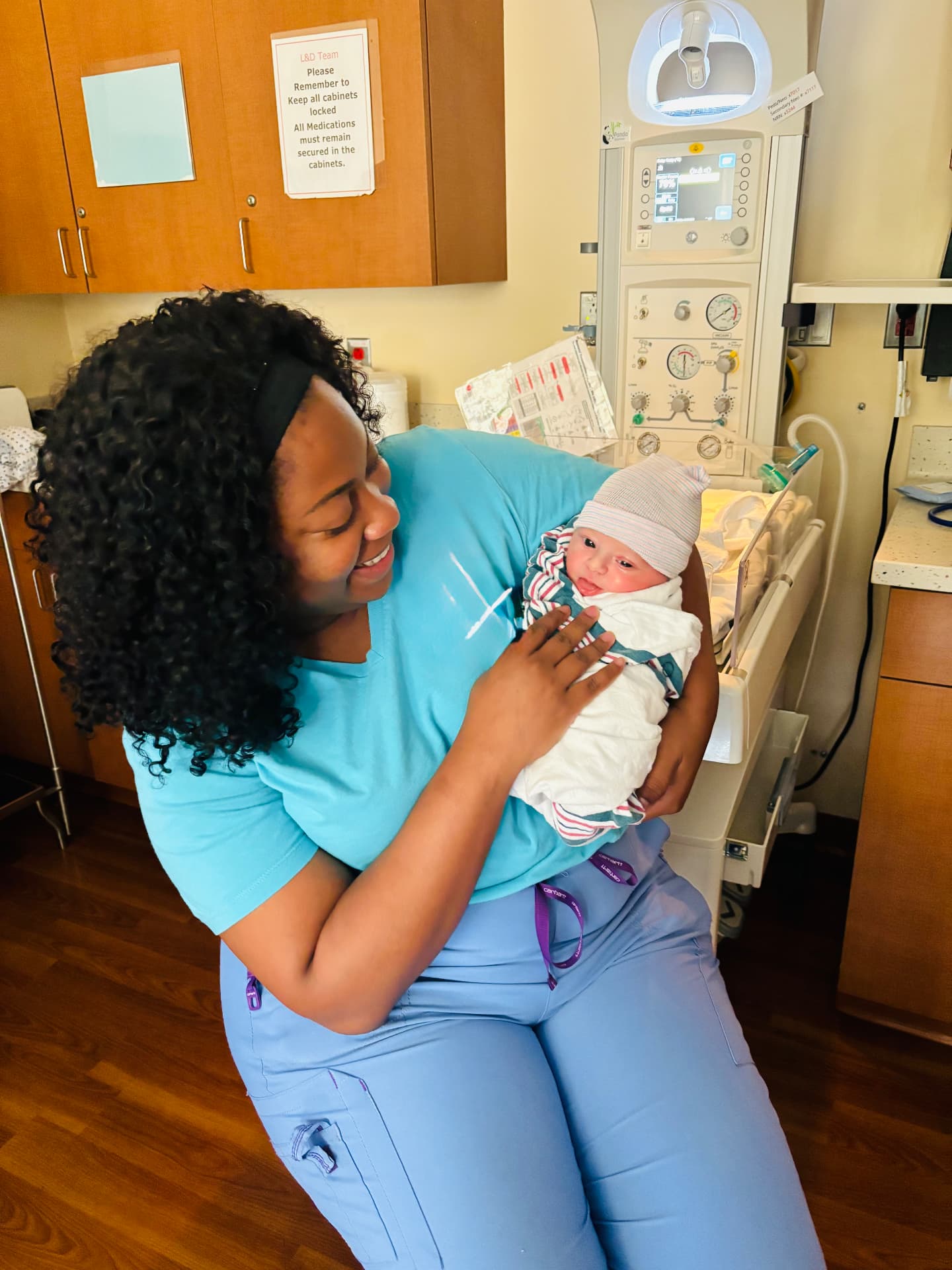 Jael in scrubs smiling down at a swaddled newborn in a hospital room