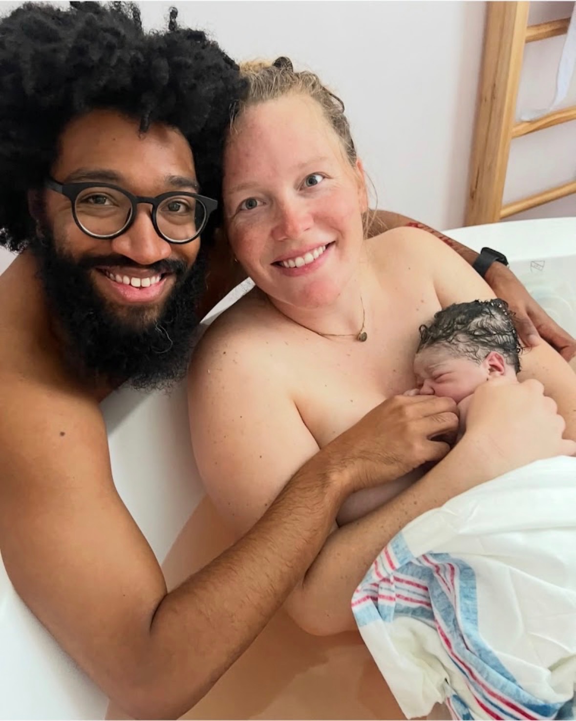 New parents smiling together in the birth tub with their newborn resting on the mother's chest